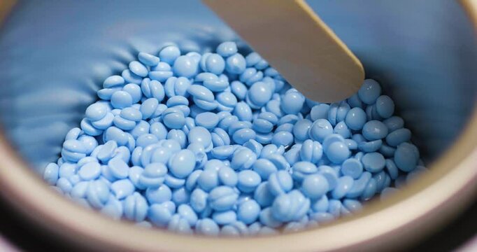 Melted light blue depilatory wax beads are being mixed with a wooden spatula in a professional wax heater, ready for a hair removal treatment