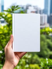 Blank Book Cover Mockup - A hand holds a blank white book cover against a blurred city and green nature background. Perfect for showcasing designs