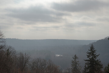 A misty valley landscape with layers of hills, trees, and a cloudy sky. The scene has a moody, overcast atmosphere with muted colors.