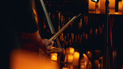 Close-up of a musician's hands with a bow playing a double bass, surrounded by lit candles. © VeNN