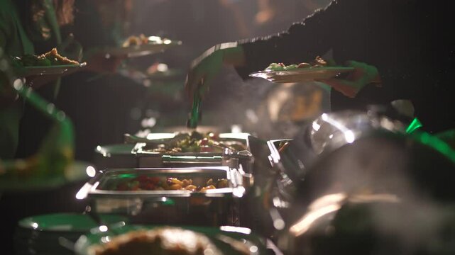 Valmiera, Latvia - November 29, 2024 - People serving themselves food at a steaming buffet table with a variety of dishes, including vegetables and meat, under warm ambient lighting