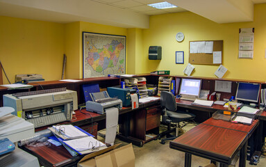 An office space is filled with desks, computers, and printers, surrounded by yellow walls. A large map hangs on one wall, and a corkboard with papers is visible.