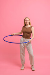 Beautiful young woman exercising with hula hoop on pink background