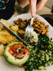First-person view: Hands skillfully using cutlery while enjoying a healthy, balanced meal featuring broccoli, avocado, potatoes, and quinoa, highlighting fresh and nutritious ingredients. 