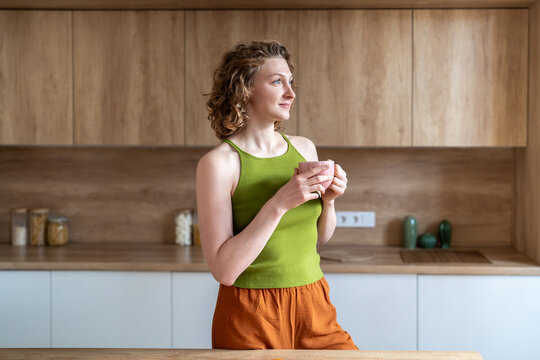 Serene woman stands in kitchen holds coffee mug takes mindful pause, reflecting on goals, affirmations. Relaxed young female in casual homewear enjoys quiet, smiles softly sets optimistic tone for day