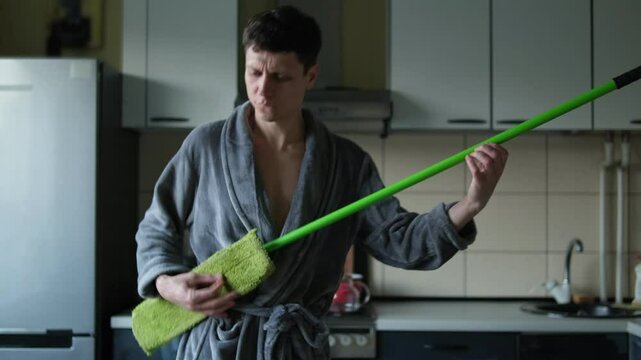 Funny guy is cleaning the room. A man in a gray bathrobe plays a mop like a rock guitar, pretends to be a famous star artist of a rock band. Male has fun like a rock musician on kitchen
