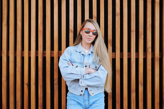 Young woman in denim jacket and jeans crossing arms while standing confidently against wooden slatted wall, showcasing casual and modern style