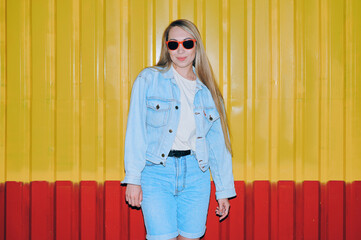Young European woman in light blue denim jacket, red sunglasses, and shorts standing in front of yellow and red vertically ridged wall, flash lighting, edited in retro style
