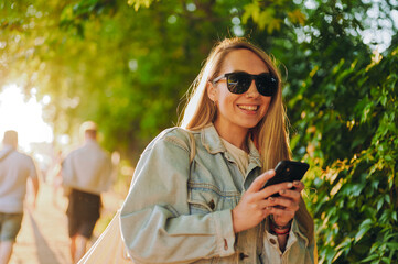 Trendy smiling woman in denim jacket actively using smartphone outdoors on sunny day capturing...