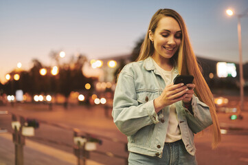 Obraz premium Young woman in denim jacket smiles softly at smartphone screen amidst glowing city nightscape, urban lights framing her natural look and modern casual style.