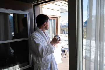 Fototapeta premium Young caucasian man wearing bath robe and drinking hot tea at his hotel room in the morning with fresh air and opened balcony Mountain View