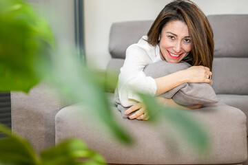 Obraz premium Happy woman with a perfect smile is lying on a comfortable sofa at home, hugging a pillow and enjoying a relaxing moment in her living room