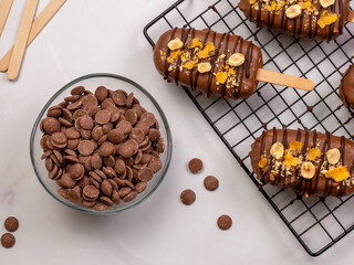 A delicious cheesecake ice cream bar on a stick, coated in chocolate and topped with nuts and Royaltyn, placed next to a transparent bowl filled with chocolate callets