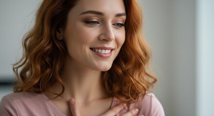 Fototapeta premium Close-up Portrait of a Happy Young Woman with Red Curly Hair, Smiling Gently While Touching Her Chest, Wearing a Pink Shirt, Soft Lighting