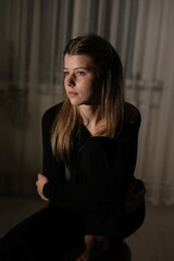 Fototapeta premium A young woman with long blonde hair sits in the dark illuminated by soft side lighting. Her gaze is thoughtful and the background is dark and blurred creating an atmosphere of melancholy and solitude.