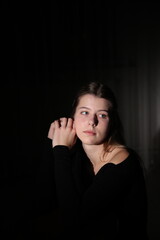 A young woman with long blonde hair sits in the dark illuminated by soft side lighting. Her gaze is thoughtful and the background is dark and blurred creating an atmosphere of melancholy and solitude.