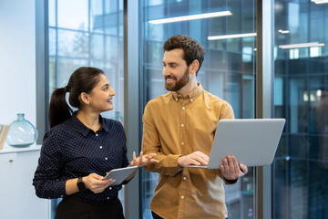 Two business colleagues, a man and a woman, collaborate using a laptop and tablet in the office.