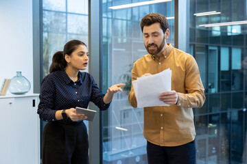 A man and woman are having a heated discussion in their office space. The man is holding papers,...