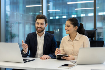Two smiling professionals are collaborating in a modern office, using laptops and sharing ideas.