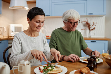 Senior couple enjoying breakfast together at home, both looking content while sharing meal at wooden table in comfortable kitchen setting, displaying mutual happiness and connection