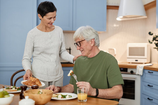 Happy elderly couple enjoying healthy breakfast in cozy modern kitchen setting. Woman serving food while man smiling and holding a fork, creating a warm atmosphere - Powered by Adobe