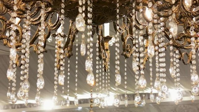 Close-up of a hanging chandelier glowing in an elegant wedding marquee, captured with aerial drone movement. Ideal for luxury interiors, high-class decor and event visuals. Cinematic drone footage.