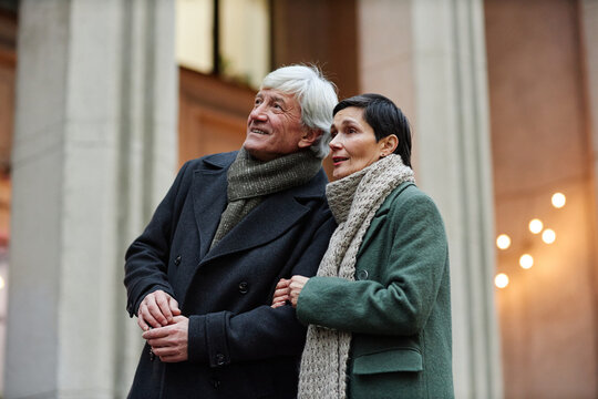 Elderly couple standing outdoors, gazing up at buildings, wearing overcoats and scarves. Signs of affection and contentment as they enjoy their surroundings