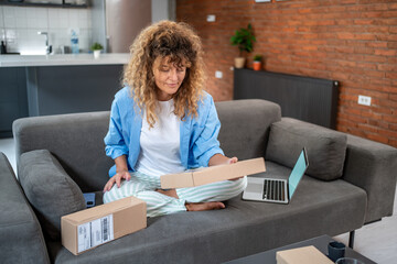 Woman opening package on sofa with laptop nearby