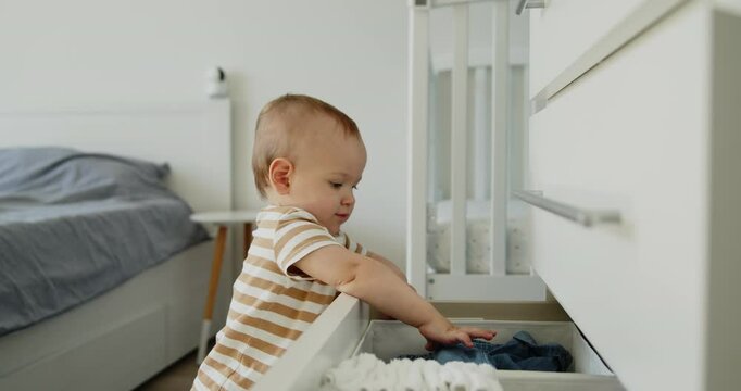 A curious baby examines the contents of a chest of drawers pulling clothes out of it. A one year old child explores the world around him. Slow motion middle shot.