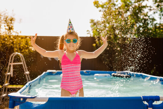 Red-haired 7-8-year-old girl celebrating her birthday in a summer pool, jumping in the water and splashing