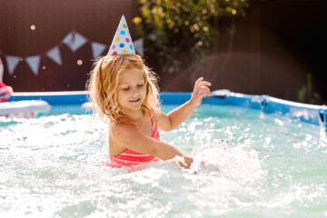 Curly haired blonde girl splashing water in the pool during a party, wearing a paper party hat