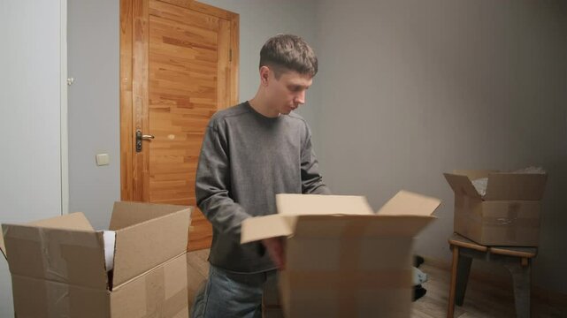 A man anxiously unpacks clothes from a cardboard box, pulling out items with frustration. The disorganized space filled with boxes suggests an unplanned move or forced relocation