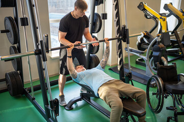 Disabled man in wheelchair train arms lifting barbell while coach demonstrate technique and support...