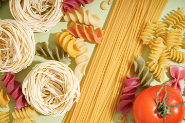 Different uncooked pasta and tomato on green background, closeup