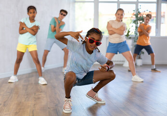 Obraz premium African boy in sun glasses performs choreographic exercises and teaches energetic mobile social dance Jazz-modern together with friends. Multi-racial group of children performs movements in studio