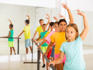 Portrait of focused tween girl practicing ballet positions near bar during group class in dance...
