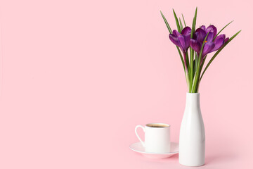 Vase with beautiful crocus flowers and cup of coffee on pink background