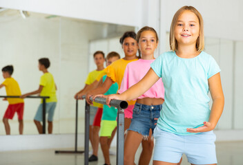 Smiling tween girl practicing classic dance moves near ballet barre during group class.