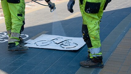 Workers in paint-stained overalls renew the marking of the bicycle path in the spring with the help of a stencil and a spray bottle