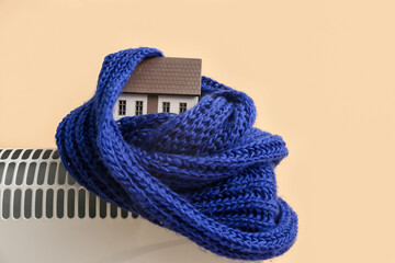 Model of house with warm scarf on electric heater against beige background. Concept of heating season