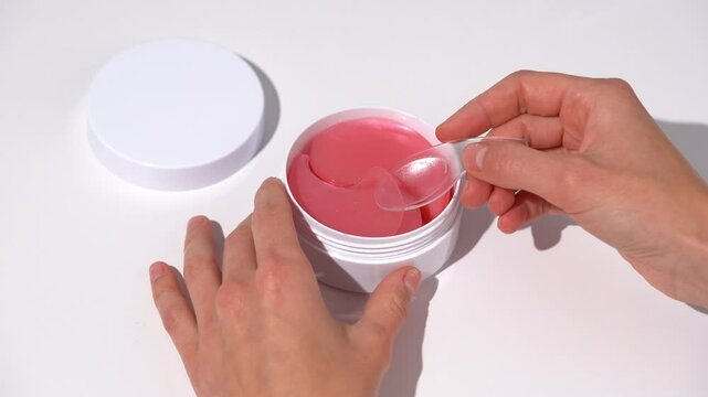 Female hands take out a pink patch from jar with spatula and demonstrate texture on white table background. Concept of beauty products under eyes for moisturizing and from dark circles under the eyes