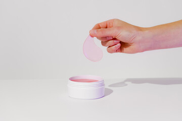 With fingers, a woman picks up a pink hydrogel patch from a mockup white jar on a white isolated...
