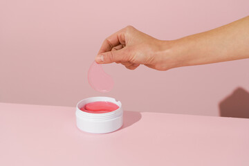 A woman’s hand holding a pink hydrogel eye patch above a white jar against a pink background. A...