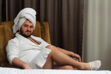 Horizontal photo. Young adult white fat man in robe with beard, towel on head, slippers on legs sits on chair in hotel bed room, looks at camera. Men day, wash, skin care, rest, spa, 8 march, check in