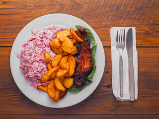 Simple meal with pork belly, potato wedges and pink coleslaw on white plate on wooden table, fork and knife on white napkin. Fine dinner with simple products.