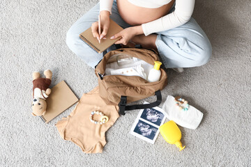 Young pregnant woman writing checklist with maternity hospital bag on carpet, top view