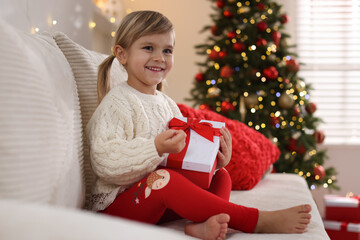 Little girl with Christmas gift box sitting on sofa at home