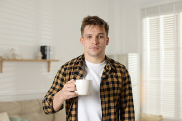 Unhappy young man with cup of coffee suffering from hangover at home