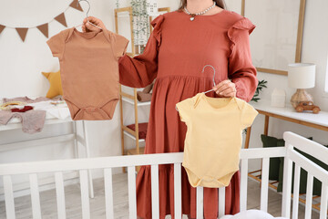 Pregnant woman with baby bodysuits near crib in bedroom