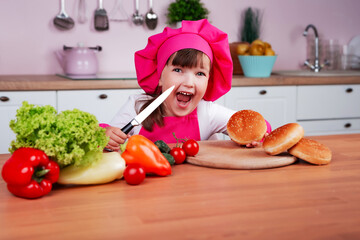 Funny happy little beautiful child girl in a chef costume prepares burgers while sitting at a table in the kitchen. Healthy eating. Child is cutting sandwich buns with a knife.	
